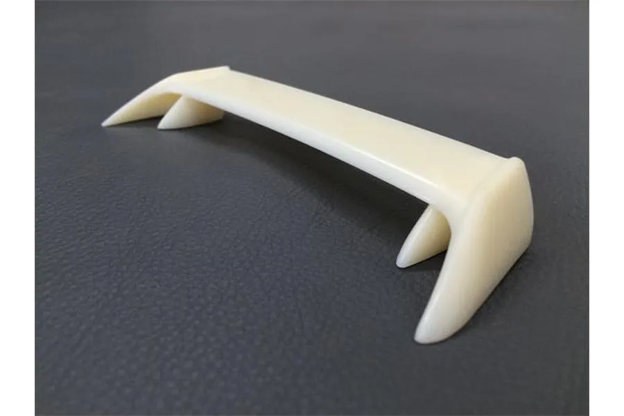 An unpainted, off-white RC car rear wing spoiler sitting on a dark grey textured surface.