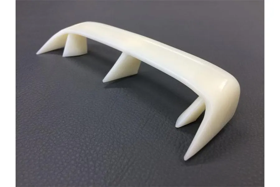An unpainted white plastic RC car rear wing spoiler with three mounting supports, viewed from a high angle against a dark grey textured background.