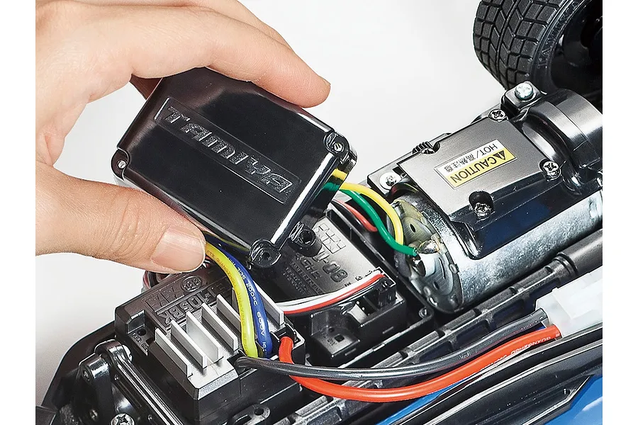 A hand holding a Tamiya branded black plastic cover over the internal electronics and motor of a TT-02 RC car chassis.