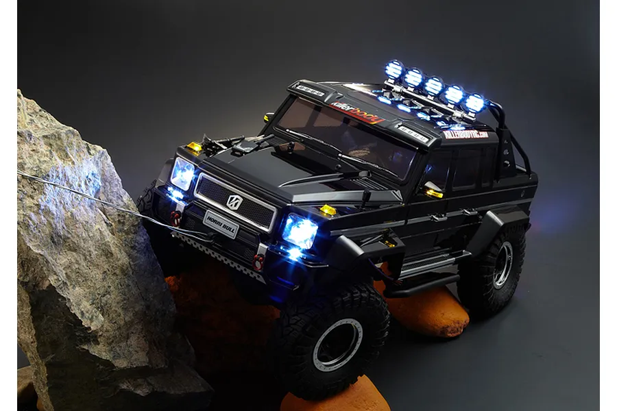 A black Killerbody Horri-Bull RC crawler body shell mounted on a chassis, climbing over a rock with active LED headlights and roof lights.