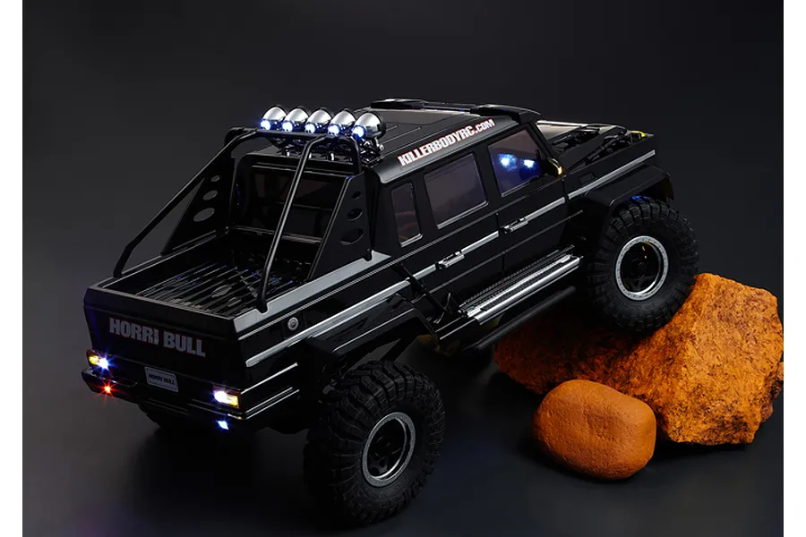 A black Killerbody Horri-Bull RC crawler body shell mounted on a chassis, posed on rocks with functional LED lights illuminated.