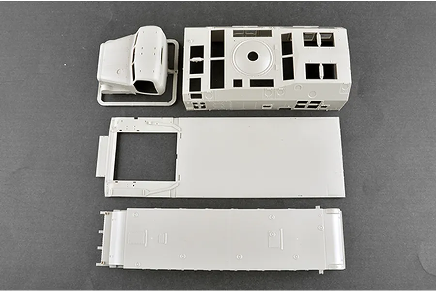 Unassembled grey plastic model kit parts for a military vehicle, including the truck cab, chassis, and body panels on a dark grey background.