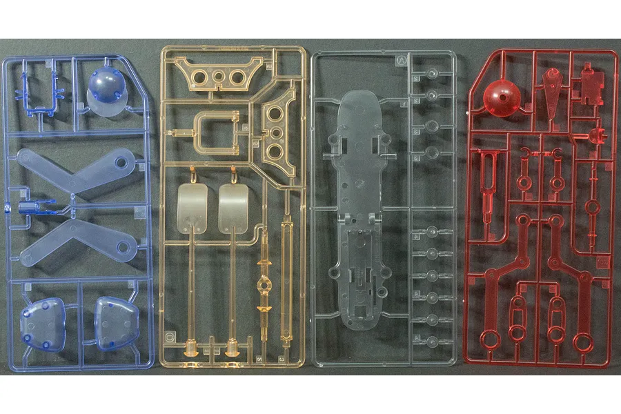 Four plastic model kit sprues in blue, orange, clear, and red, laid out on a dark grey surface.