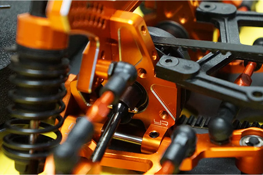 Close-up of an orange anodized aluminium RC car suspension assembly with black plastic components and a drive belt.