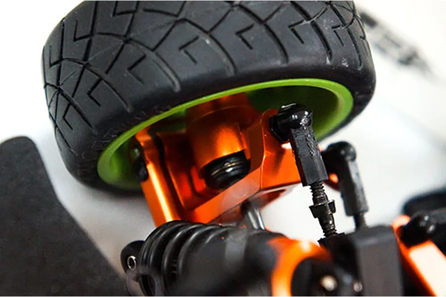 Close-up of an orange anodized aluminium RC steering arm installed on a vehicle with a black tyre and green wheel rim.