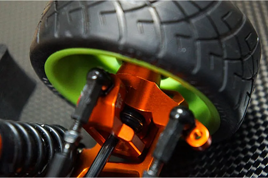 Close-up of an orange anodized aluminium RC steering hub installed on a vehicle with a green wheel rim and black tire.