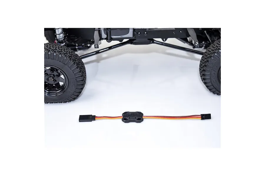 Castle Creations B-Link Bluetooth adapter module with wiring harness laid on a white surface in front of an RC vehicle chassis.