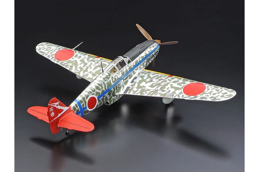 A high-angle studio shot of a 1/48 scale Tamiya Kawasaki Ki-61 Hien model aircraft with a camouflage pattern and red tail fin.