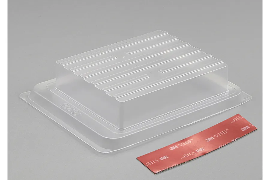 A clear plastic RC monster truck bed accessory with a strip of 3M VHB double-sided adhesive tape on a grey background.