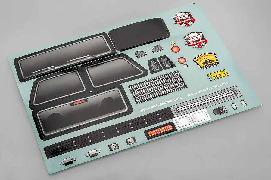A sheet of RGT RC crawler decals featuring window tints, grille, lights, and branding stickers on a light blue backing.