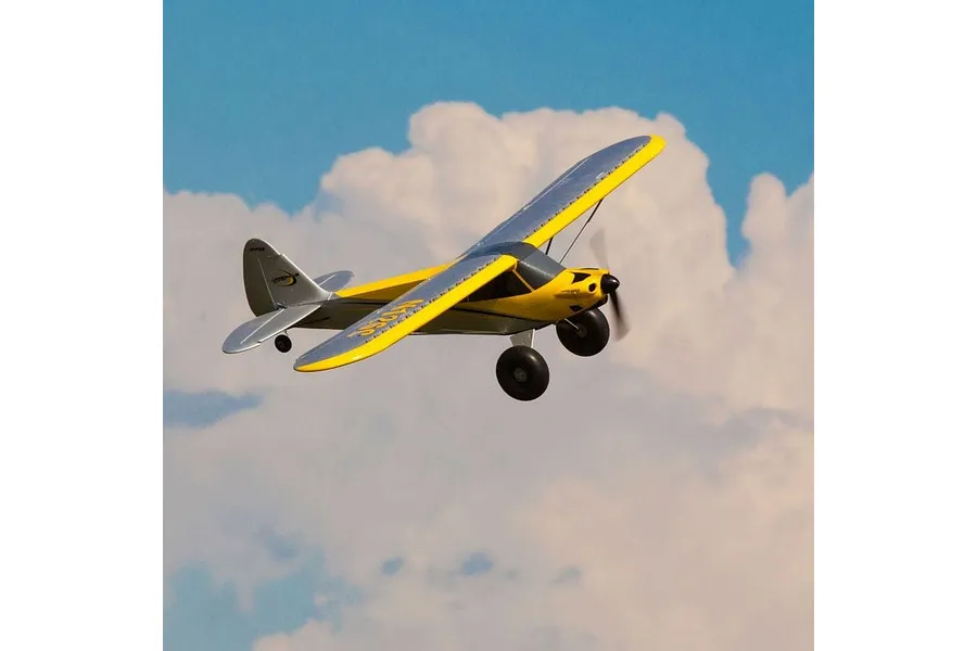 A yellow and silver Carbon Cub S2 RC plane flying against a blue sky with white clouds.