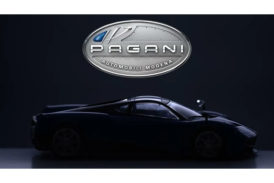 A dark silhouette of a Pagani Huayra model car positioned below the silver Pagani Automobili logo against a dark background.