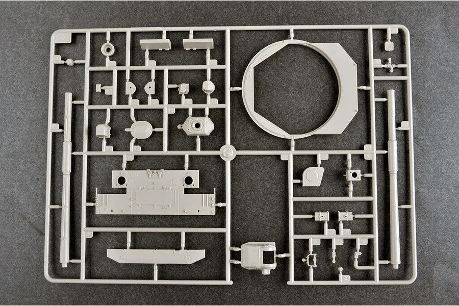 A grey plastic model kit sprue containing various tank parts, including a large turret ring and gun barrels, laid out on a dark grey surface.