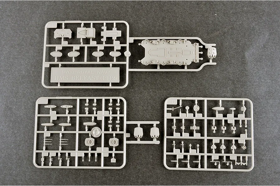 Three grey plastic model kit sprues for a 1/700 scale HMS Belfast ship model, showing various small parts and a deck section on a dark grey surface.