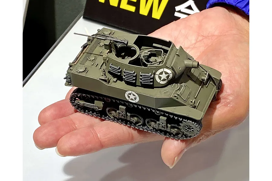 A hand holding a small, assembled 1/48 scale Tamiya M8 Howitzer Motor Carriage model tank painted in olive drab with US military star markings.