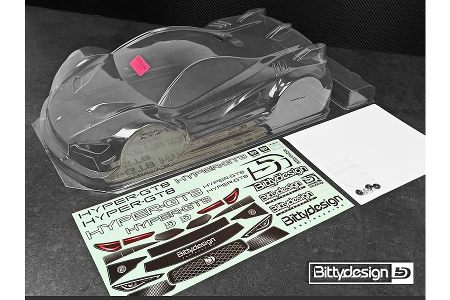 Unpainted clear polycarbonate 1/8 scale GT body shell for RC cars, including a decal sheet, window masks, and mounting hardware on a dark surface.