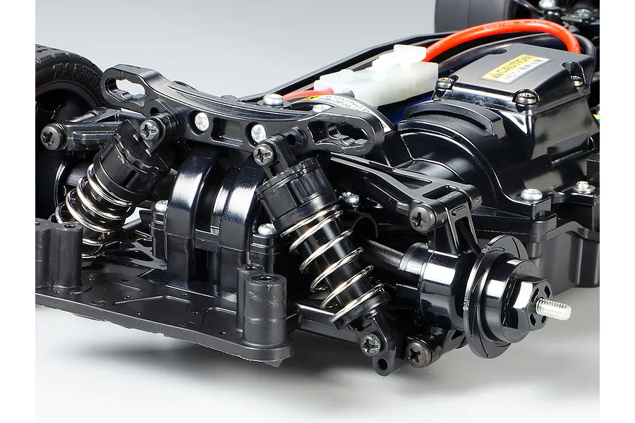 Close-up of the front suspension and chassis of a Tamiya TT-02 RC car kit, showing black plastic arms, coil-over shocks, and wheel hubs.