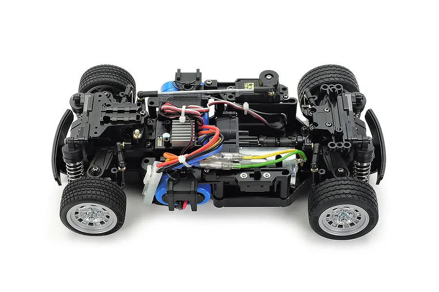Top-down view of a Tamiya MB-01 RC car chassis showing the motor, suspension, wheels, and internal electronics on a white background.