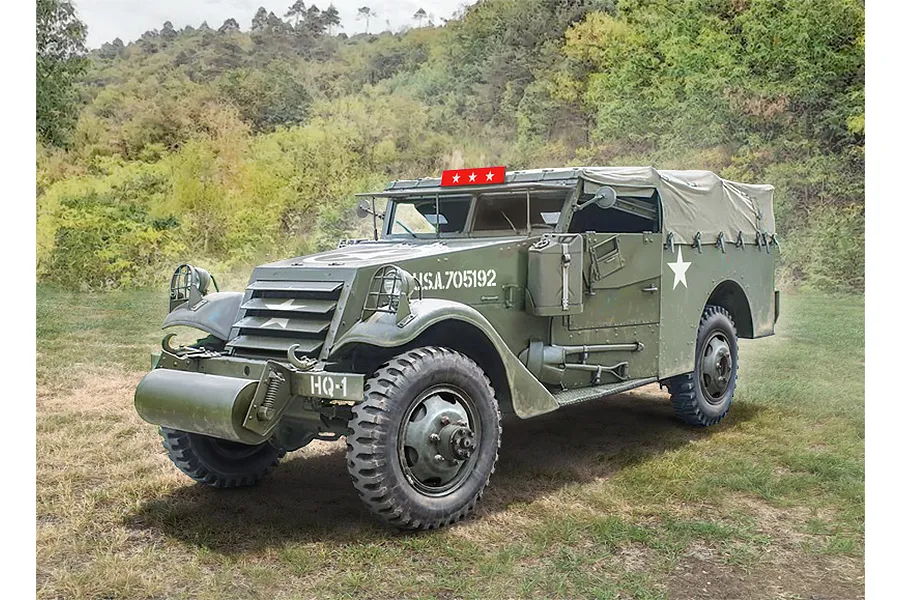 A scale model of an olive drab M3A1 Scout Car parked on a grassy field with a forest background.