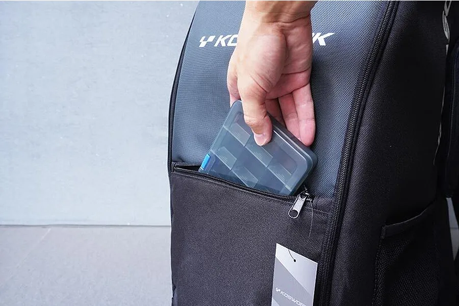 A hand placing a small, grey plastic parts organizer box into the zippered front pocket of a black Koswork RC crawler backpack.