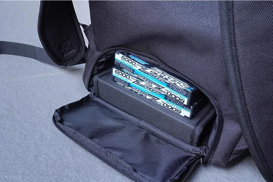 A close-up view of a black Koswork crawler backpack with a zippered compartment open, revealing two LiPo batteries stored inside.
