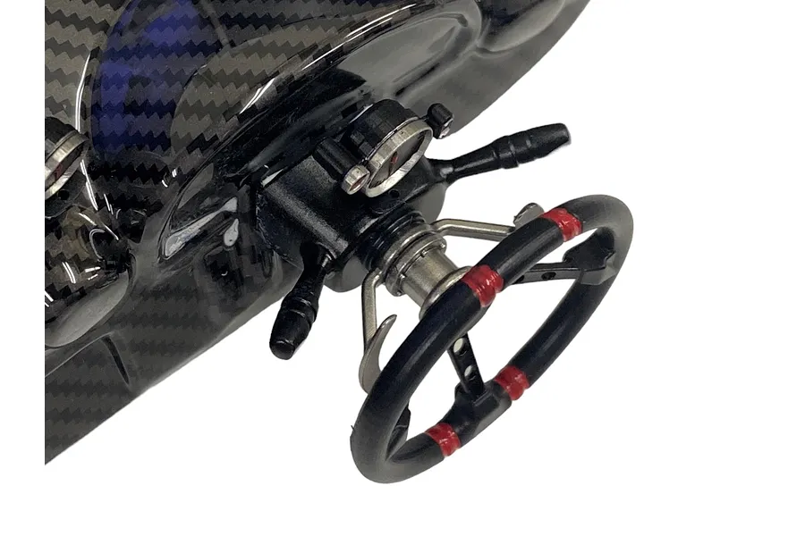 A close-up of a scale model racing steering wheel attached to a carbon fiber dashboard, featuring black and red detailing.