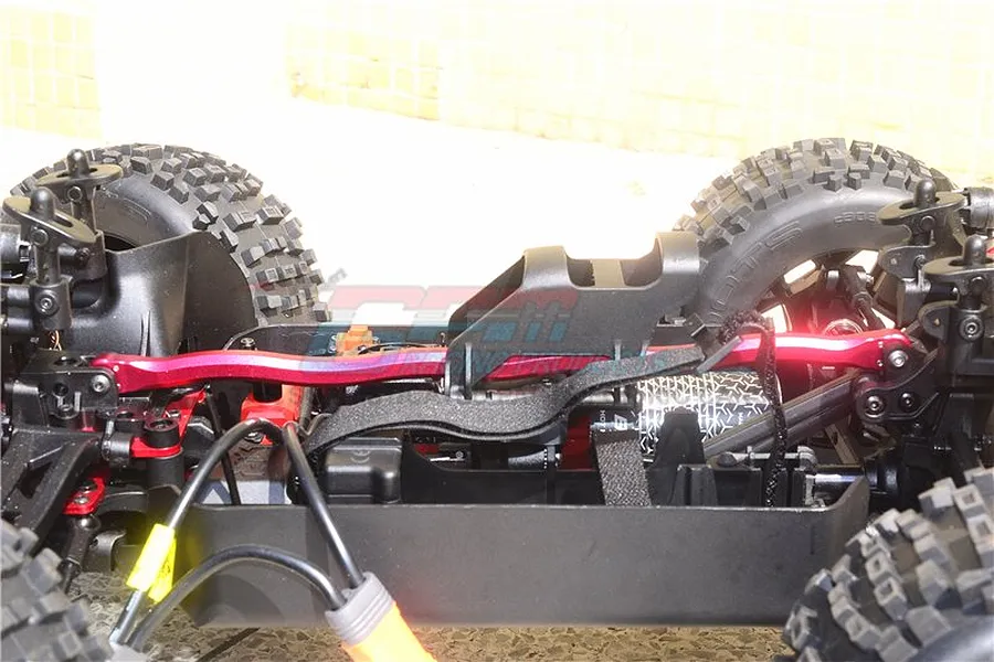 A red aluminium centre brace bar installed on an RC car chassis, showing the central structural component between the front and rear suspension towers.