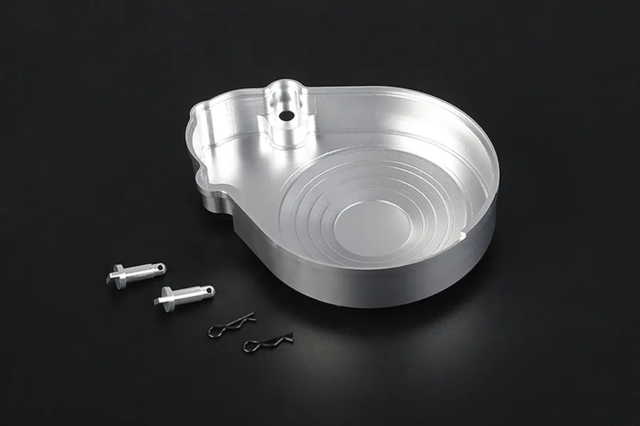 A silver aluminium RC transmission case component with two small metal pins and two black R-clips on a dark background.