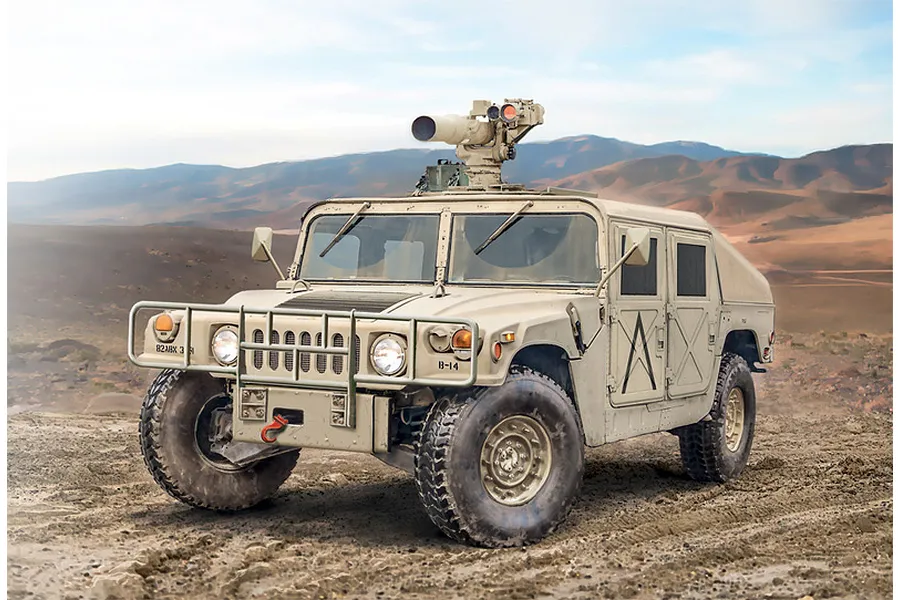A tan-colored HMMWV M966 TOW carrier military vehicle parked on a dirt surface with a desert mountain landscape in the background.