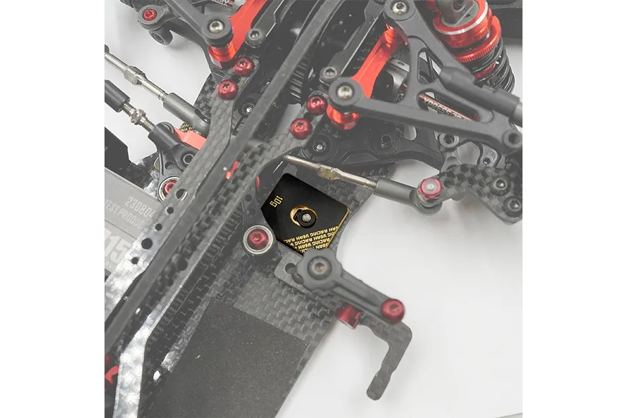 A close-up view of a 10g Yeah Racing black brass chassis balancing weight installed on a carbon fiber RC car chassis.