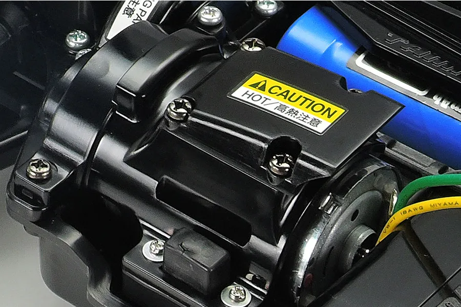 Close-up of the motor and gear cover assembly on a Tamiya TT-02B RC buggy chassis, featuring a yellow caution label.