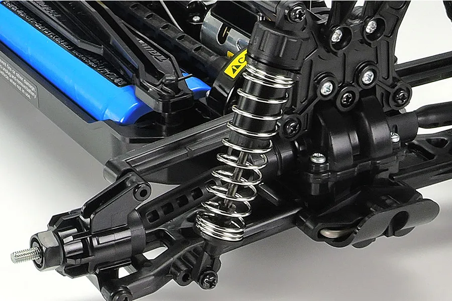 Close-up of the rear suspension and chassis of a Tamiya TT-02B RC buggy, showing the coil-over shock, black plastic suspension arms, and a blue battery pack.