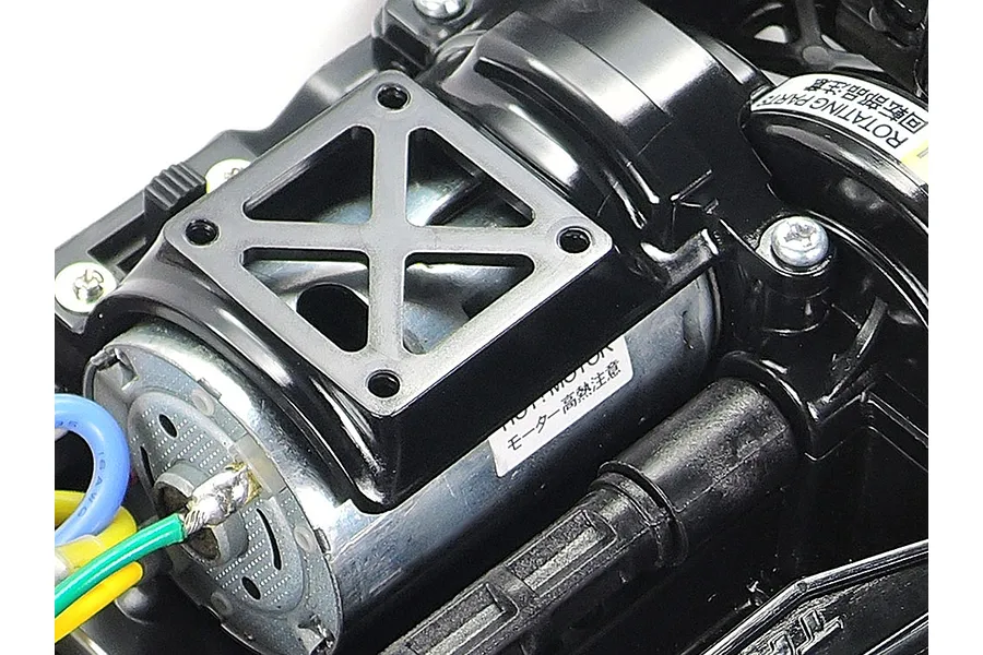 Close-up of a brushed electric motor installed in a Tamiya TT-02 RC car chassis, featuring a black motor mount and warning labels.