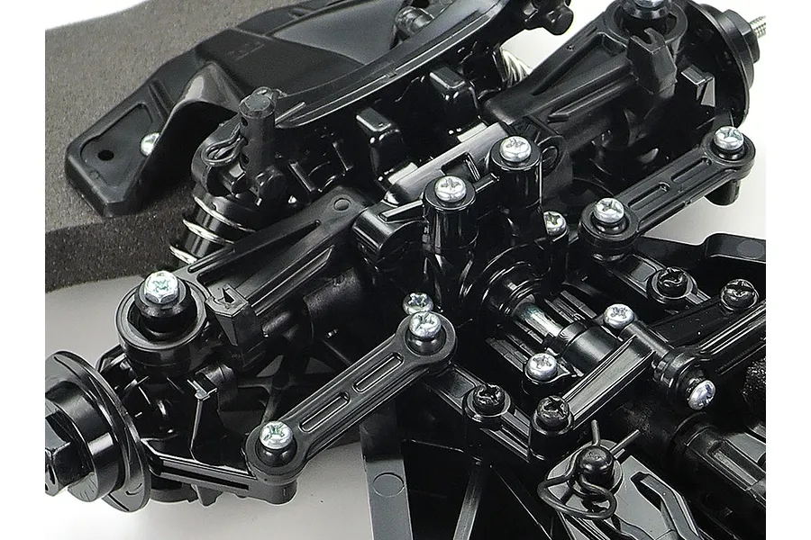 Close-up of the black plastic front suspension assembly of a Tamiya TT-02 RC car chassis, showing steering linkages, shock towers, and screws.