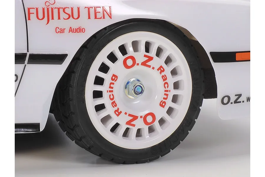 Close-up of the front wheel and fender of a white Tamiya Toyota GT-Four ST165 RC rally car, featuring a white O.Z. Racing wheel and Fujitsu Ten branding.
