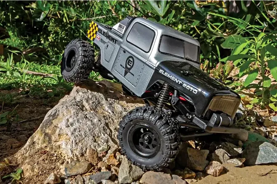 Element RC Enduro24 Ecto 1/24 scale RC rock crawler in silver and black, climbing over a rock in an outdoor garden setting.