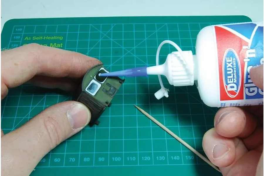 A person applying Deluxe Materials Glue 'N' Glaze to a small scale model window on a green self-healing cutting mat.