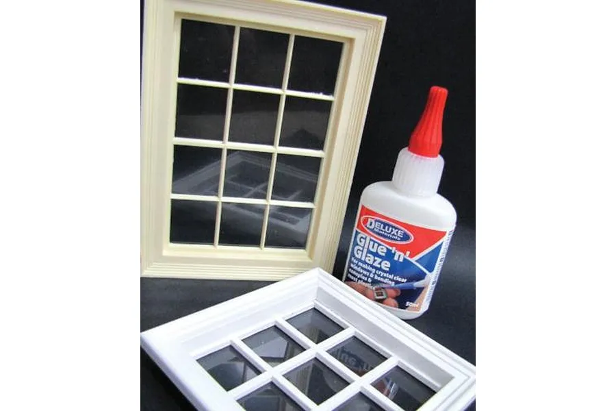 A bottle of Deluxe Materials Glue 'N' Glaze adhesive next to two miniature model window frames on a black background.