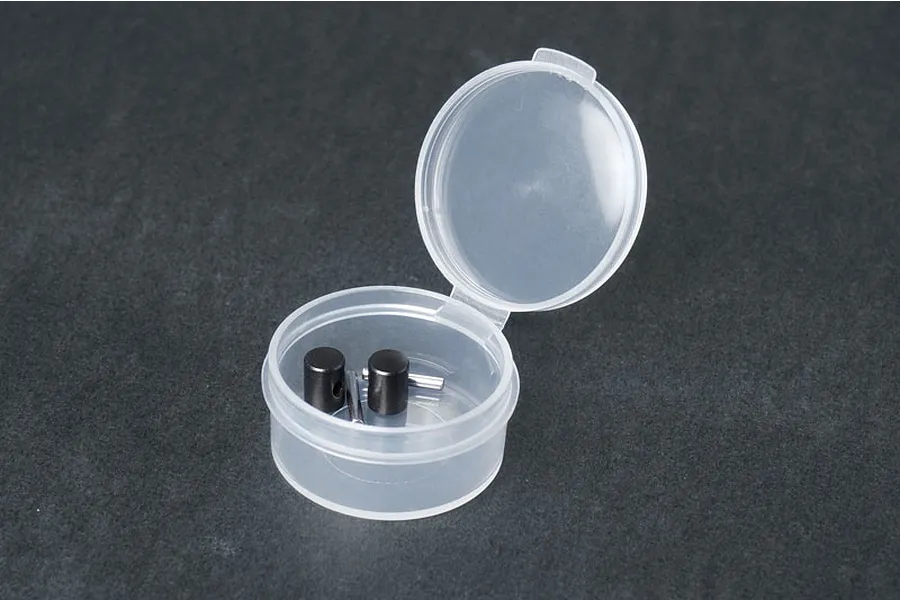 Koswork CVA coupler and pin rebuild kit components inside a small, open, translucent plastic storage container on a dark grey surface.