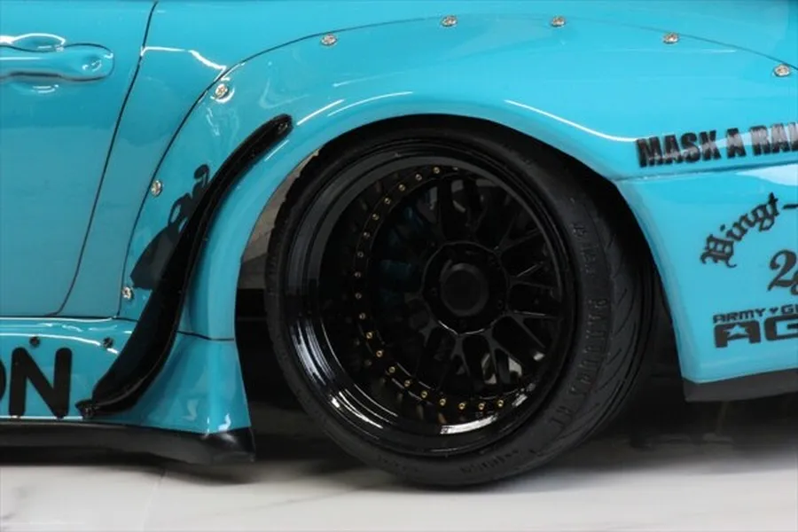 Close-up of a light blue RC car body with a wide fender flare and a black multi-spoke deep-dish wheel with gold lug details.