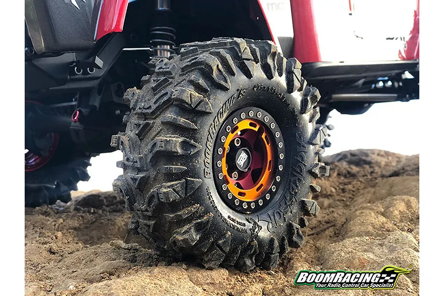 A close-up of a Boom Racing 1.9 inch Aggressor tyre mounted on a red RC crawler vehicle, driving over rocky terrain.