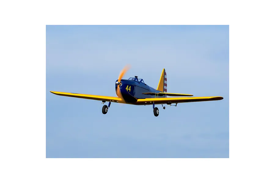 A Hangar 9 PT-19 RC plane in flight against a clear blue sky, featuring a dark blue and yellow livery with the number 44 on the side.