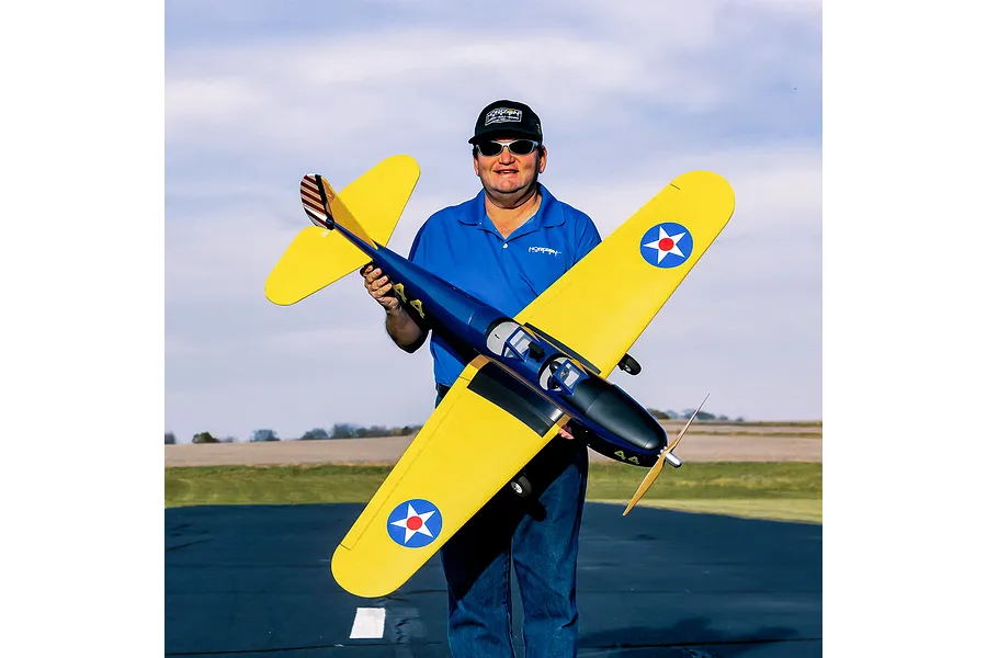 A man holding a yellow and blue Hangar 9 PT-19 RC plane outdoors at an airfield.