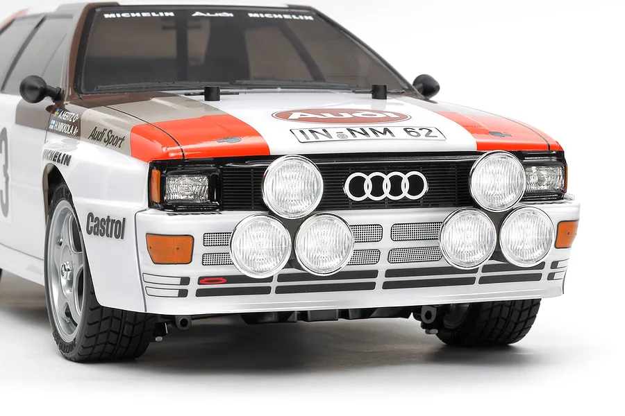 Front three-quarter view of a Tamiya Audi Quattro A2 RC car with rally livery, featuring prominent round auxiliary driving lights on the front grille.