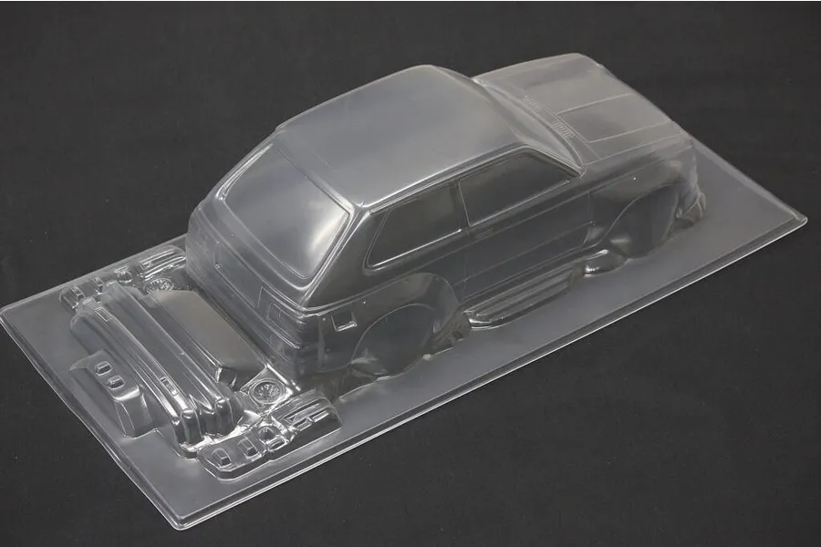 A clear, unpainted 1/10 scale Toyota Starlet KP61 RC car body shell with separate bumper and accessory parts on a black background.