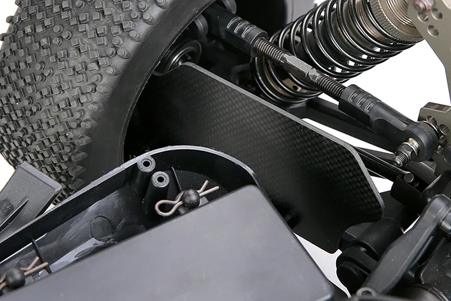 Close-up of a carbon fibre suspension arm guard installed on an RC vehicle chassis, showing the weave pattern and surrounding suspension components.