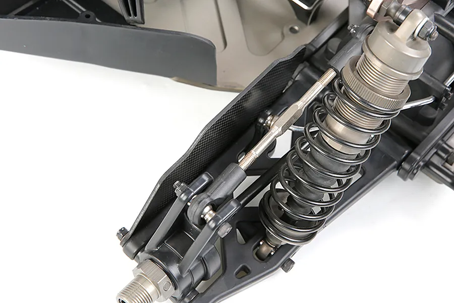 A close-up view of a carbon fibre rear suspension arm guard installed on an RC car chassis, featuring a coil-over shock absorber and suspension linkage.