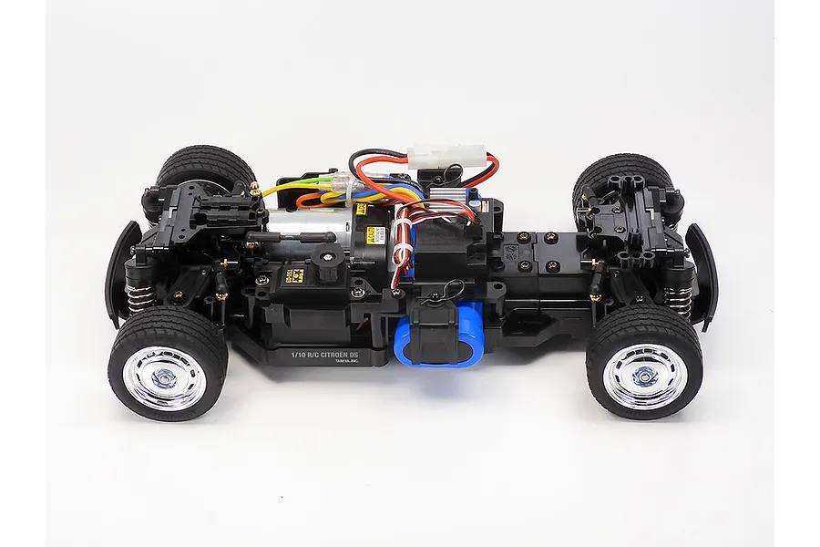 A high-angle studio shot of the Tamiya MB-01 Citroen DS RC car chassis, showing the motor, battery, and suspension components on a white background.