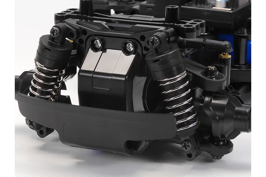 Close-up of the front suspension and bumper assembly of a Tamiya MB-01 RC car chassis, featuring black plastic parts and silver coil springs.