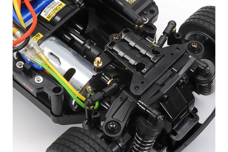 Close-up of a Tamiya MB-01 RC car chassis showing the electric motor, suspension components, and wiring on a white background.